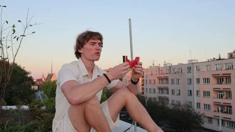 Young man sitting on rooftop at sunset eating last slice of watermelon while war Stock Footage 315413843