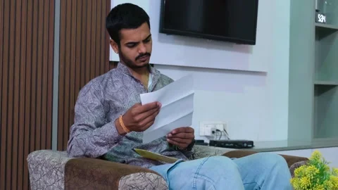 Young man sitting on a sofa and carefully reading a letter or document at home Video stock 330390766