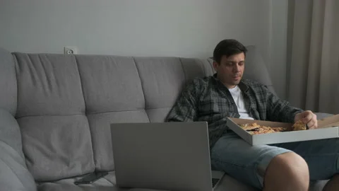 Young man is sitting on sofa eating slice of pizza and watching video on laptop. Stock Footage 167790061
