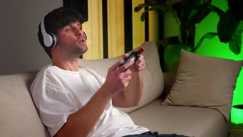 Young man is sitting on the sofa, holding a joystick in the living room 스톡 동영상 235923843