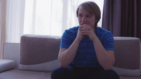 Young man sitting on the sofa at home in the living room waiting for something Видео 73060983