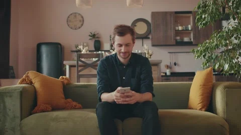 A young man is sitting on the sofa at home using a smartphone Stock Footage 138535814