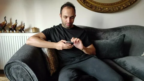 Young Man Sitting on Sofa is Looking at his Phone Stock Footage 144045593