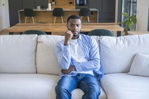 Young man sitting on a sofa in a modern living room, looking thoughtful and.. Stock Photos