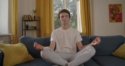 Young man sitting at sofa practicing yoga relaxation after busy workweek slow Stock Footage 143515887