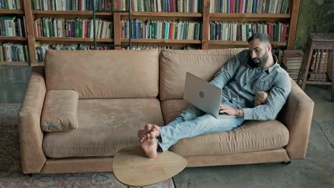 Young Man Sitting On Sofa In Relaxed Position With Laptop And Talks On The Phone Video stock 140225184