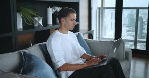Young man sitting on sofa, studying or working on laptop from home. Male student Stock Footage 250588644
