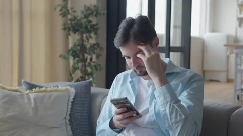 Young Man Sitting On Sofa Stunned By Unexpected News On His Smartphone Stockbeeldmateriaal 301242947