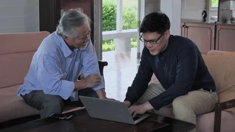 Young man sitting on sofa talking and teach adult man about use laptop in liv 스톡 동영상 137880946