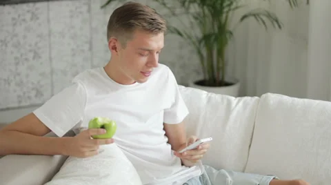 Young man sitting on sofa using cellphone and eating apple Stock Footage 27691910