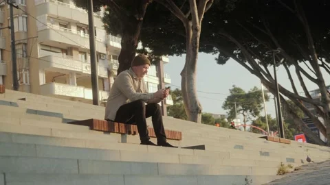 A young man is sitting on the steps outside at sunrise and texting on the phone. Stock Footage 221147114