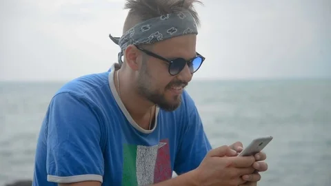 Young man is sitting on a stone and using smartphone near the sea. Portrait of Stock Footage 79405654