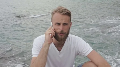 Young man is sitting on a stone and using smartphone near the sea. Portrait of Stock Footage 81038498