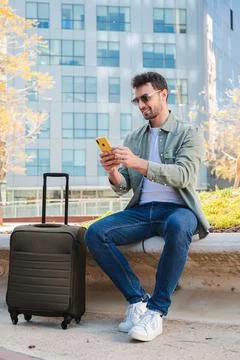 Young man sitting with suitcase while checking messages on smartphone outdoors 스톡 사진