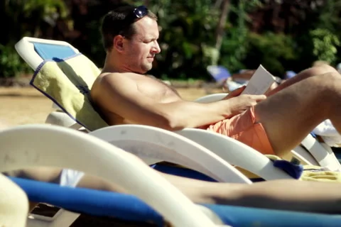 Young man sitting on sunbed and reading book by the pool   Stock Footage 1068705