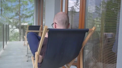 Young man sitting in a sunbed on the balcony and using mobile phone. Video stock 62519940