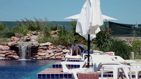 Young man sitting on sunbed under umbrella by the pool Stock Footage 69784405