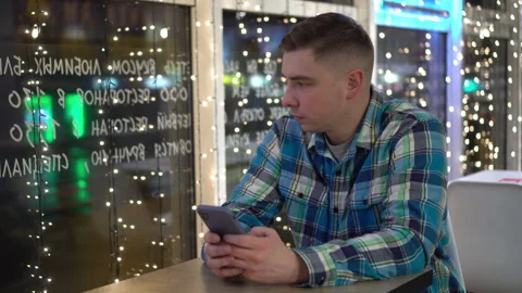 A young man is sitting at a table in a cafe in the evening and texting on the Stock-Footage 166678462