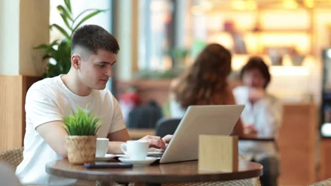 Young man is sitting at a table in a cafe and working at a laptop. Guy designer Stock Footage 312632876