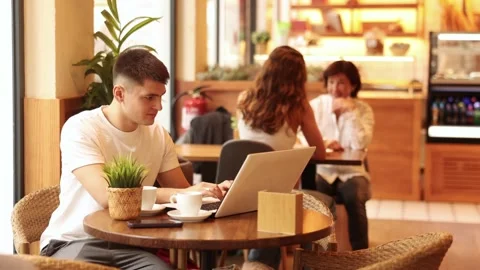 Young man is sitting at a table in a cafe and working at a laptop. Guy designer Stock Footage 319990520