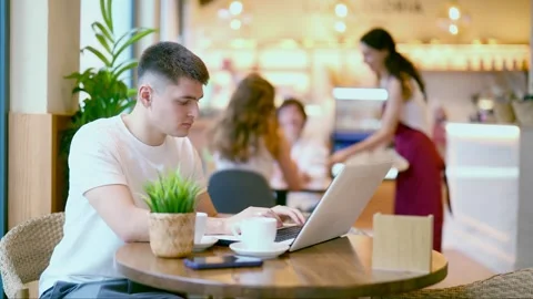 Young man is sitting at a table in a cafe and working at a laptop. Guy designer Stock Footage 335616520