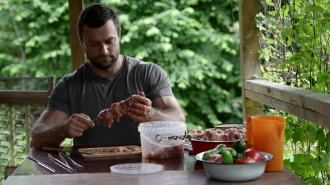 A young man is sitting at a table in a gazebo and stringing pieces of raw meat Stock Footage 163661546