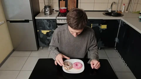 Young man sitting at a table, hesitating between two donuts, smelling and Stock Footage 305554292