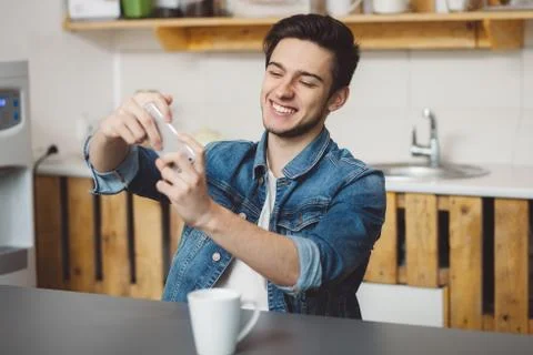 Young man sitting at a table in the kitchen with his mobile phone Stock Photos