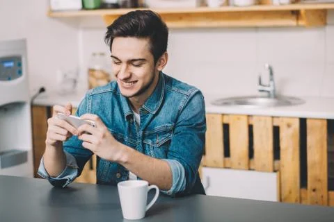 Young man sitting at a table in the kitchen with his mobile phone Stock Photos