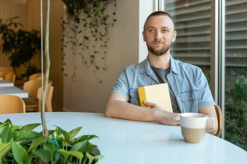 Young man sitting at a table in a modern space holding a book and a cup dur.. Stock Photos