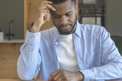 Young man sitting at a table in a modern kitchen, looking thoughtful while .. Stock Photos