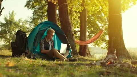 Young man is sitting in the tent at sunset and using laptop on hiking in the Stock Footage 98671834