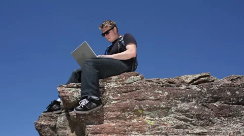 Young man sitting at top of cliff with laptop Stock Footage 61657218