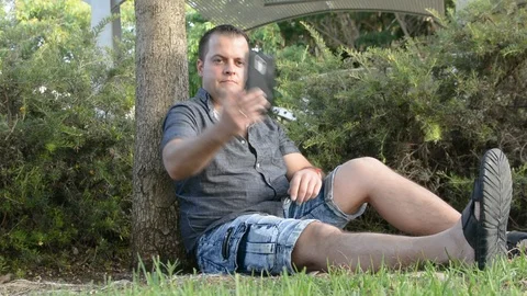Young man sitting by the tree and talking on mobile phone Видео 91432416