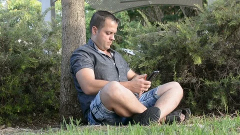 Young man sitting by the tree and talking on mobile phone Vidéo 91434171