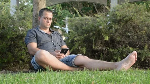 Young man sitting by the tree and talking on mobile phone Vídeo Stock 91448233