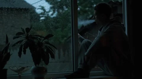 Young man sitting on window sill at rainy evening, smoking in silence. Moody Stock Footage 311449183