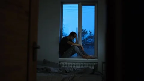 Young man sitting on a windowsill in a dimly lit room, holding his head in distr Stock Footage 296274475