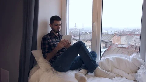Young man is sitting on a windowsill of a large window, overlooking his Stock Footage 70528837