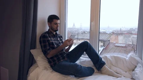 Young man is sitting on windowsill, reading from a tablet and enjoys city Stock Footage 70528794