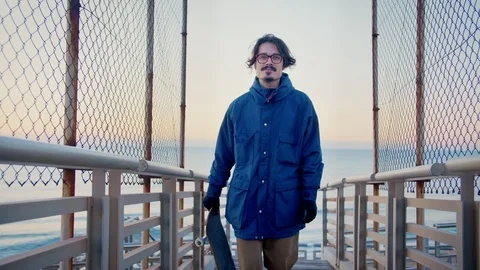 Young man skater walking by small bridge in beach in cold morning with skate Stock Footage 100860809