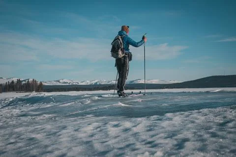 Young man with skis and backpack on frozen lake with mountain view Stock Photos