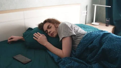 A young man sleeps in bed, then turns off the alarm and continues to sleep Stockbeeldmateriaal 196813519