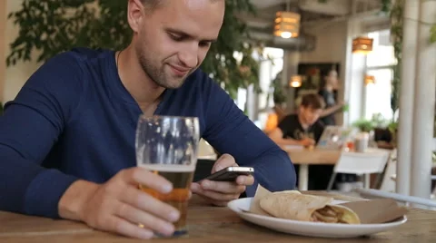 Young man with smartphone drinking beer in cafe Stock Footage
