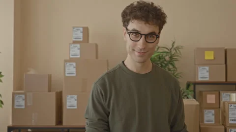 Young man smiles at camera amid stacked shipping boxes and parcels on shelv.. Stock Footage 337435585