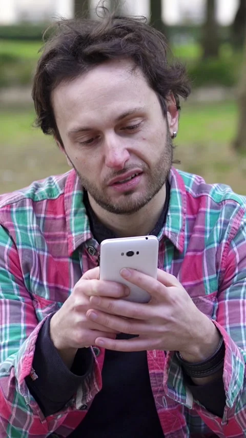 Young Man Smiles While Texting on Smartphone in Park Stock Footage 304972491