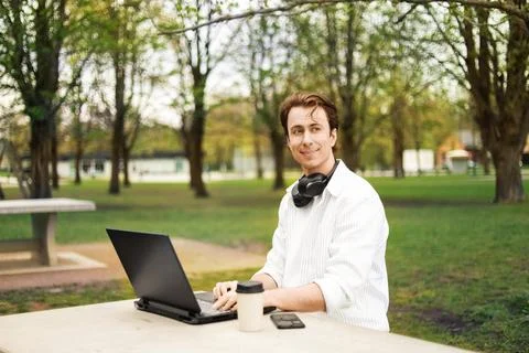 A young man smiles while working on a laptop at a stone table in a park. He.. Stock Photos