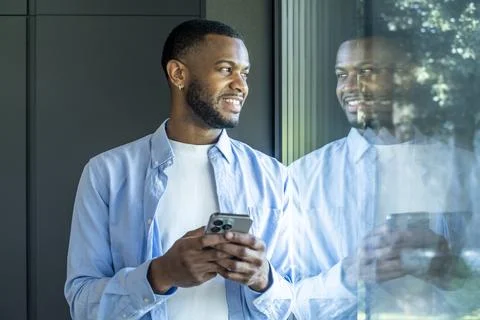 Young man smiling and looking out a window while holding a smartphone, refl.. Stock Photos