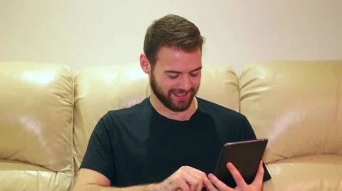Young man smiling and touching screen on his iPad mini at home 스톡 동영상 59956217