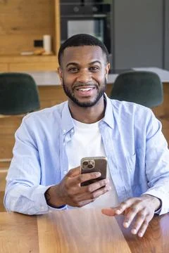 Young man smiling at camera while using smartphone at home kitchen table, c.. Fotos Stock